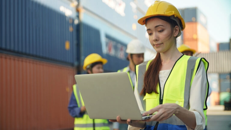 Eine Arbeiterin mit Helm und Warnweste steht vor Frachtcontainern und überprüft mit dem CPC-Tracker auf ihrem Laptop den Transport der Industriegüter.