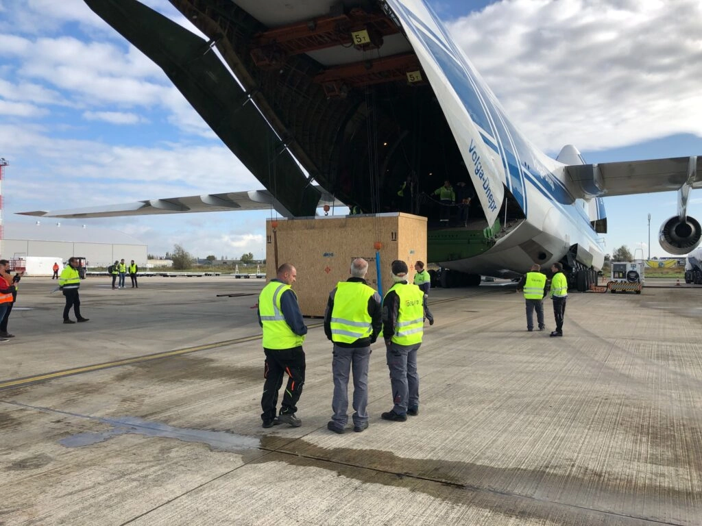 Männer in Warnweste überwachen die Ladung einer Ware in einer Transportkiste, die in ein Flugzeug gehoben wird.