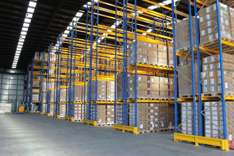 Viele gelb-blaue Hochregale nebeneinander in einer Lagerhalle, in denen zahlreiche Kartons lagern.