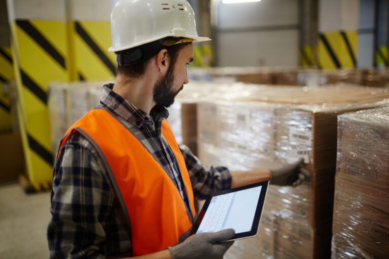 Lagermitarbeiter vergleicht mit einem Tablett die Angaben auf einer Transportverpackung innerhalb der Lagermanagement-Software