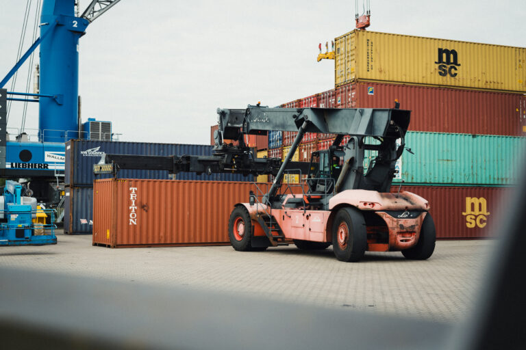 Ein orangener Frachtcontainer, der vor anderen gestapelten Containern gelagert ist, wird durch einen speziellen Kran angehoben.
