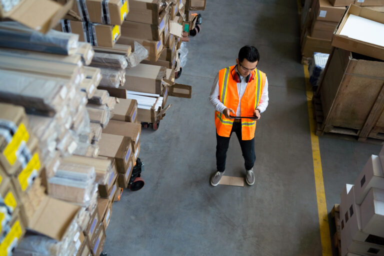 Ein Lagermitarbeiter steht zwischen einem Hochregal mit verpackten Gütern und anderen Transportkisten aus Holz und Pappe und überprüft auf einem I-Pad mit der Supply-Chain-Management-Software die Lieferkette.